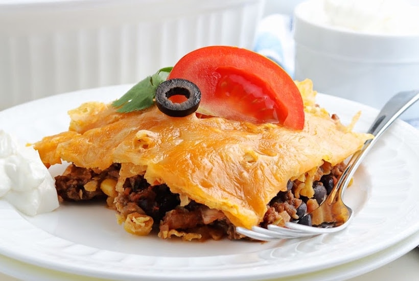 A slice of walking taco bake garnished with chopped tomato and black olive on a white dish with a fork.