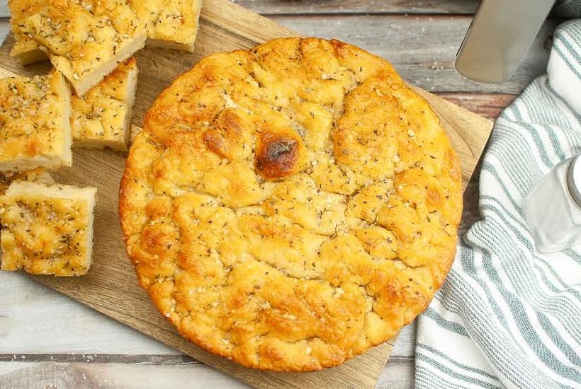 Overhead view of a round of homemade focaccia on a cutting board, with sliced bread on the left.