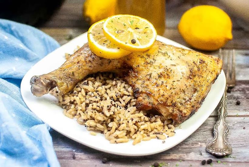 White plate filled with an air fried chicken leg quarter with brown rice and garnished with lemon slices.
