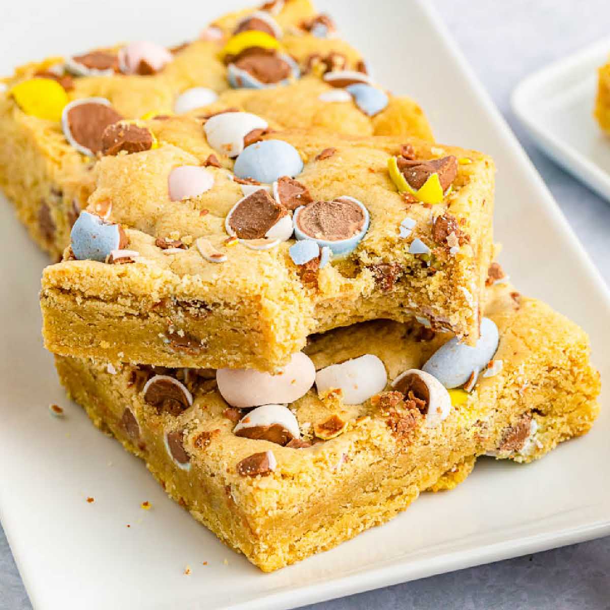 Close up of Cadbury Mini Egg Cookie Bars stacked on a white serving tray with a bite taken from the top piece.