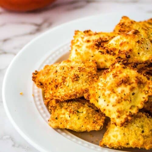 Closeup view of air fryer ravioli on a white plate with a bowl of marinara sauce.