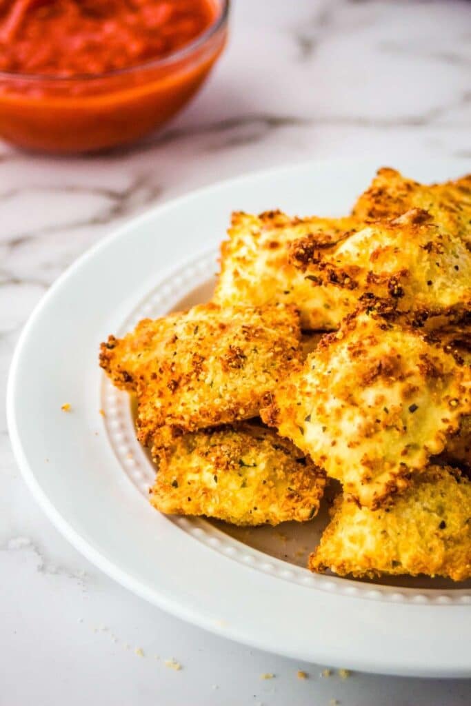 Closeup view of air fryer ravioli on a white plate with a bowl of marinara sauce.