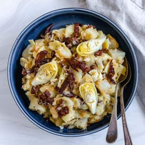 Potato salad with artichokes and sun dried tomatoes in a blue serving bowl with spoons.
