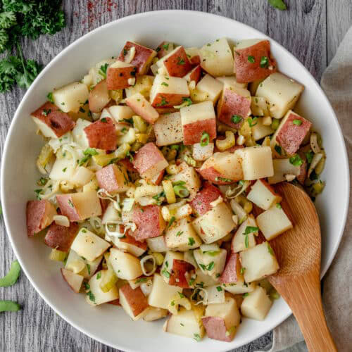 Chopped red potatoes with celery and green onions with a mustard dressing in a white serving bowl with a wooden spoon.