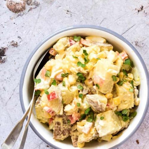 Creamy red potato salad with red peppers and garnished with chopped chives in a white serving bowl.