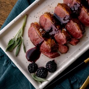 Slices of cooked duck breast topped with blackberry and sage sauce on a white platter.