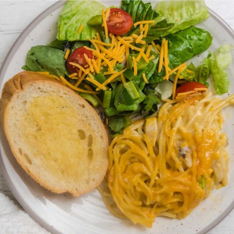 White dinner plate filled with cheesy chicken spaghetti, green salad with tomato and a slice of garlic bread.