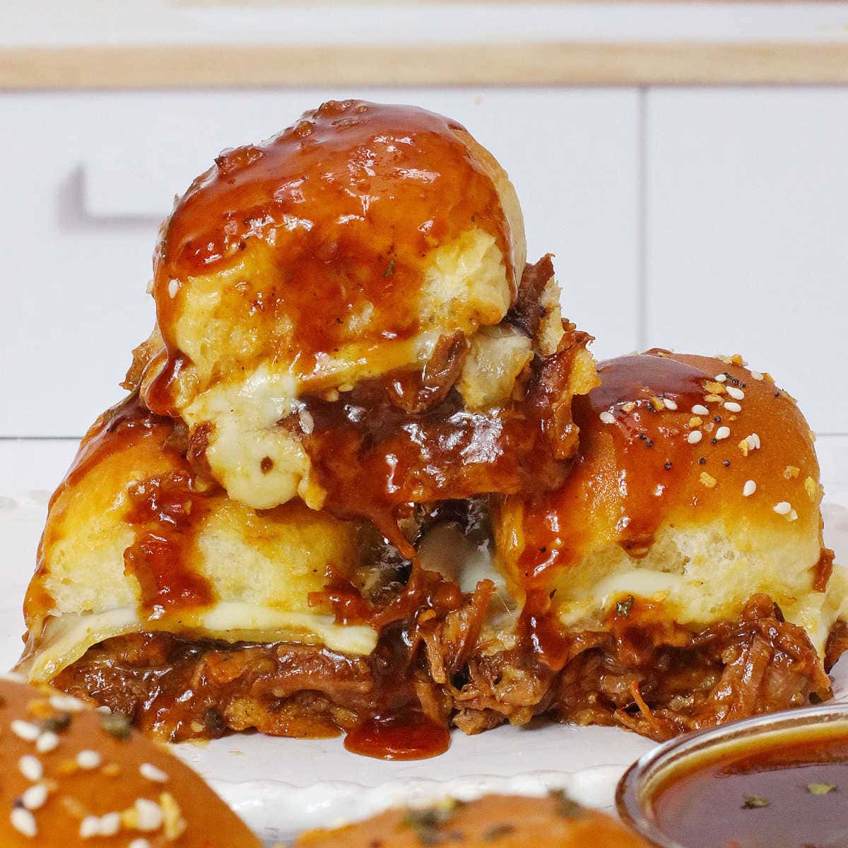 A stack of three beef brisket sliders with cheese and barbecue sauce on a white plate.