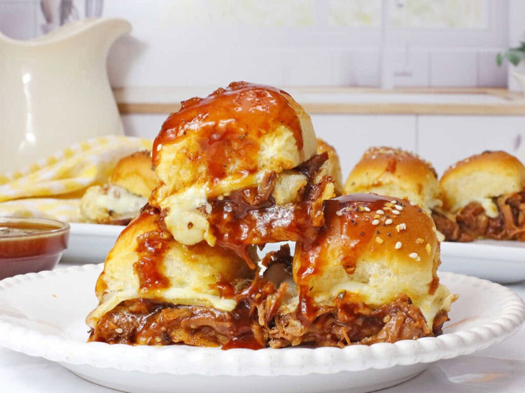 Three leftover brisket sliders with barbecue sauce on a white plate with more sandwiches in the background.