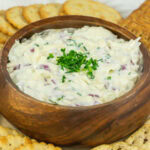 Closeup view of wooden bowl filled with homemade Jarlsberg cheese dip garnished with chopped chives and surrounded by crackers.