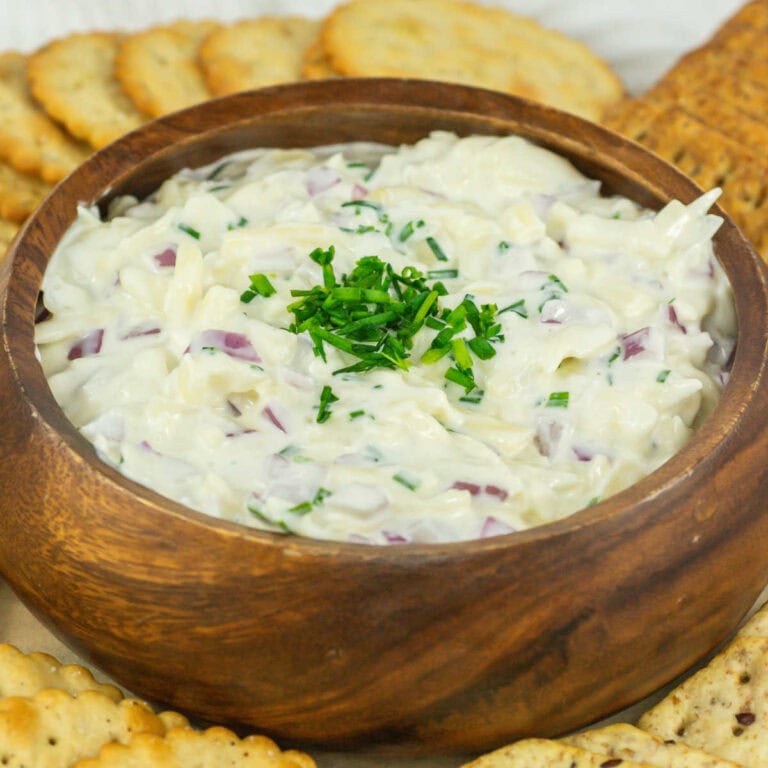 Closeup view of wooden bowl filled with homemade Jarlsberg cheese dip garnished with chopped chives and surrounded by crackers.