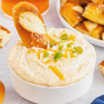 A white bowl filled with ranch beer cheese dip garnished with onions, shredded cheese and a soft pretzel.
