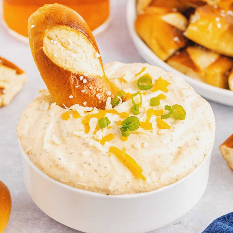 A white bowl filled with ranch beer cheese dip garnished with onions, shredded cheese and a soft pretzel.