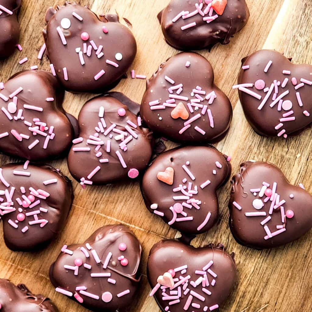 Homemade chocolate covered peanut butter hearts with pink sprinkles on a wooden cutting board.
