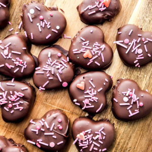 Homemade chocolate covered peanut butter hearts with pink sprinkles on a wooden cutting board.