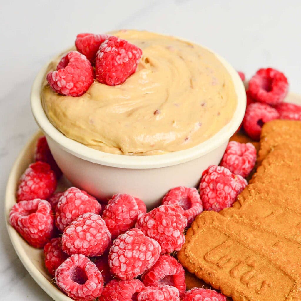 Small bowl of cookie butter dip on a plate with raspberries and Lotus cookies.