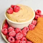 Small bowl of cookie butter dip on a plate with raspberries and Lotus cookies.