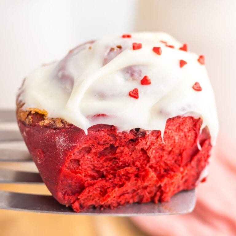 Closeup view of a red velvet cinnamon roll with cream cheese frosting and heart shaped sprinkles on a spatula.