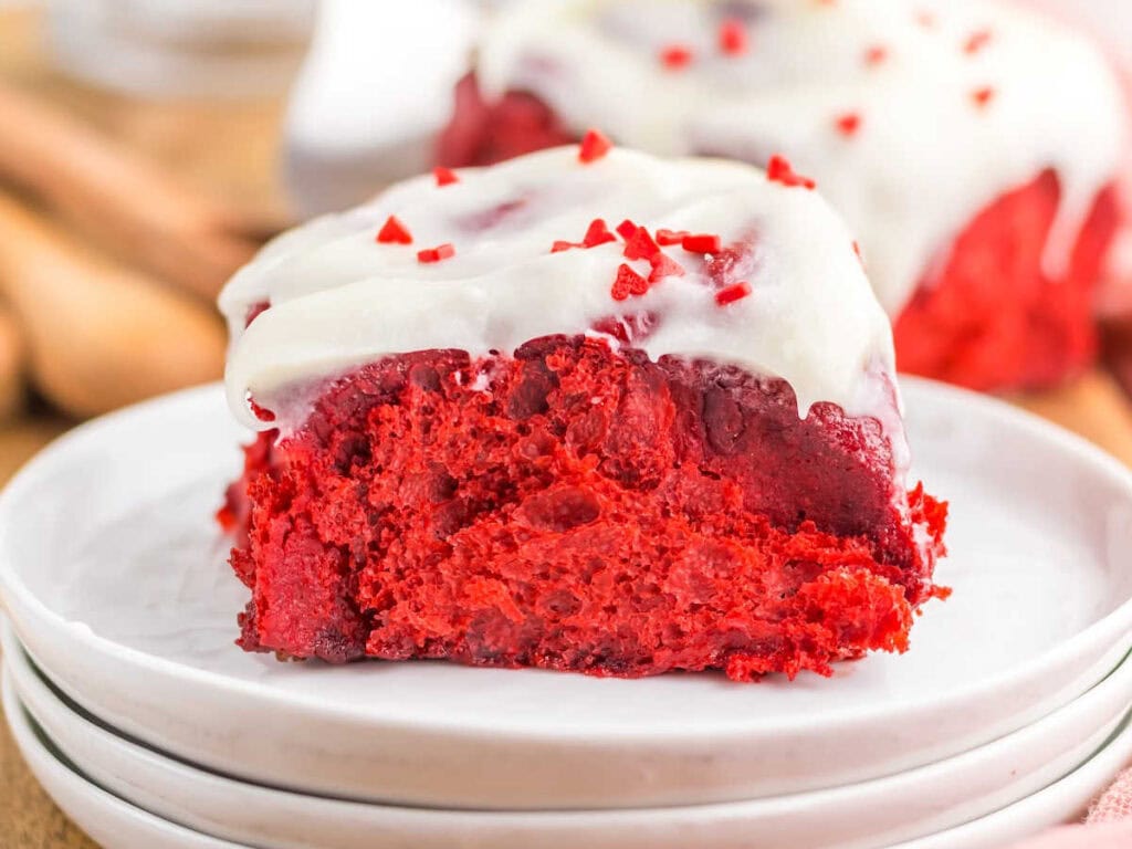 Side view of a red velvet cinnamon roll topped with cream cheese frosting and heart shaped sprinkles on a stack of white plates.