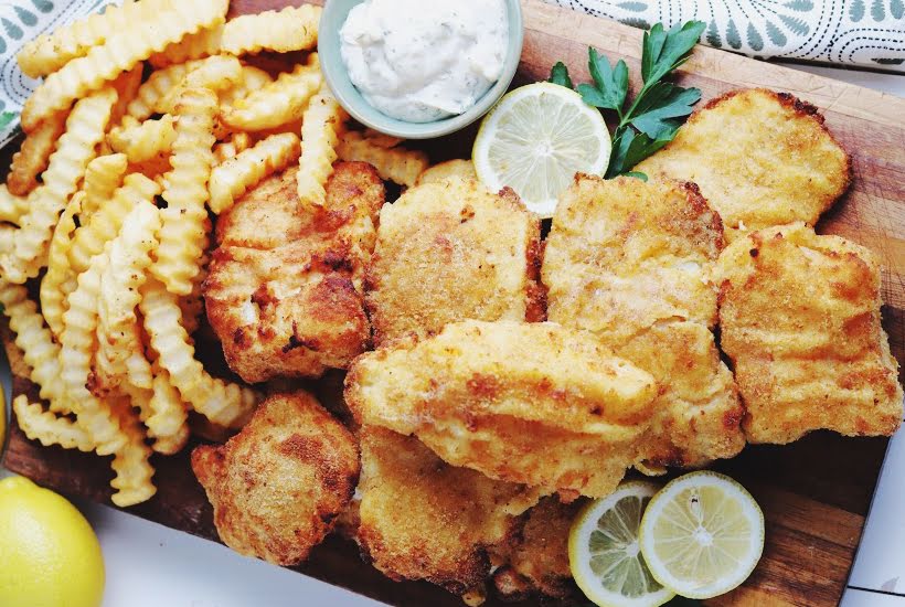 Pieces of air fried cod and French fries on a wood cutting board with a bowl of tartar sauce and lemon slices.