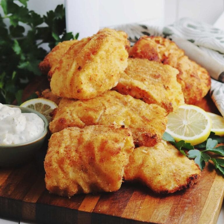 Breaded cod pieces stacked on a wood board with lemon slices and a bowl of tartar dipping sauce.