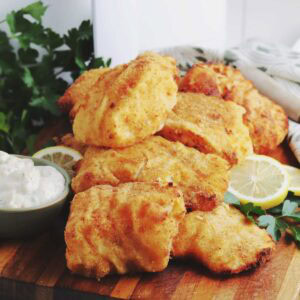 Breaded cod fish cooked in the air fryer stacked on a wood board with a bowl of tartar sauce and lemon slices.