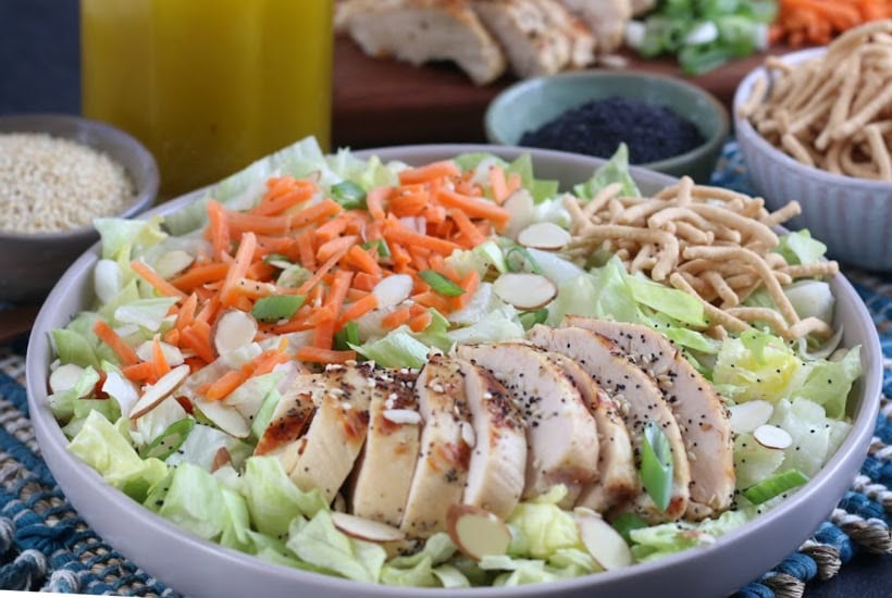 Gray bowl filled with Chinese chicken salad garnished with sliced almonds, crunchy noodles, green onion and shredded carrots.