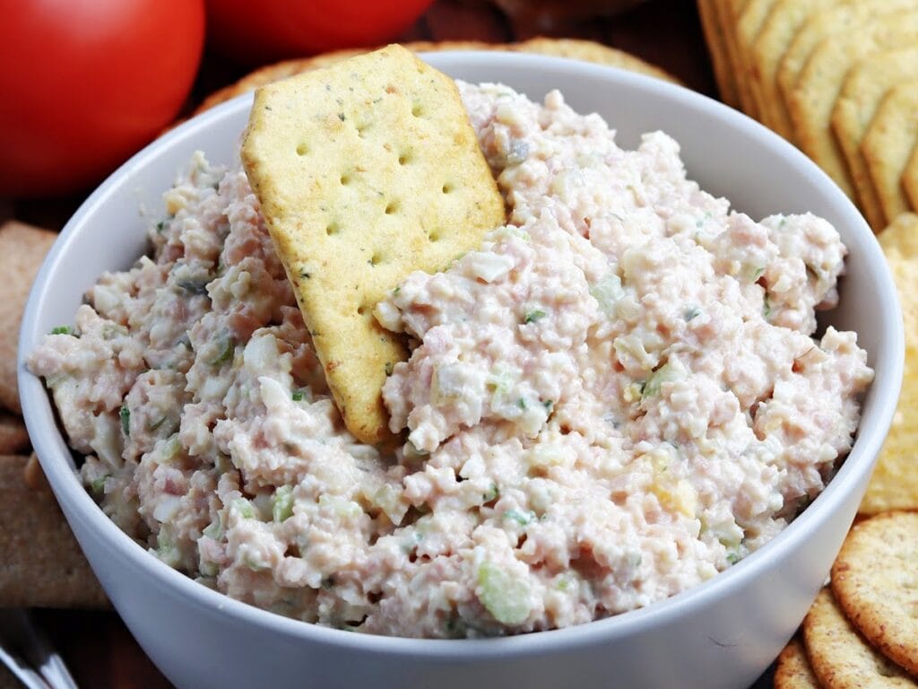 White bowl of ham salad spread with a cracker dipped in it.