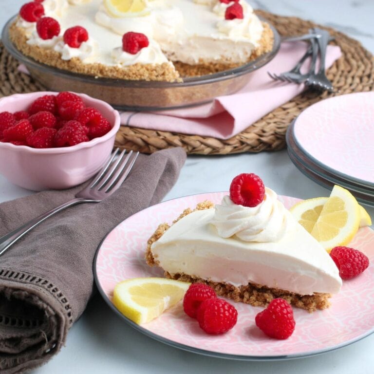 Slice of lemonade pie garnished with lemon slices and raspberries on a pink plate with partial pie in the background.
