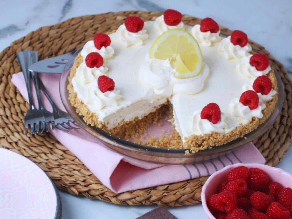 Old fashioned lemonade pie with a slice removed and garnished with whipped cream, raspberries and lemon slices.