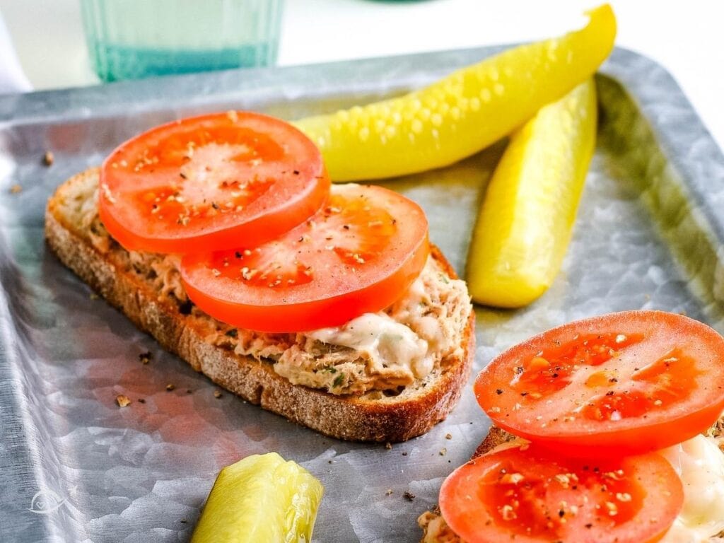 Two open faced tuna melts with tomato slices and pickle spears on a metal serving tray.