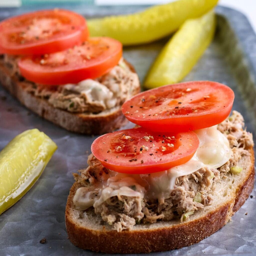 Closeup view of open faced tuna melt sandwiches with tomato slices and pickle spears on a metal serving tray.