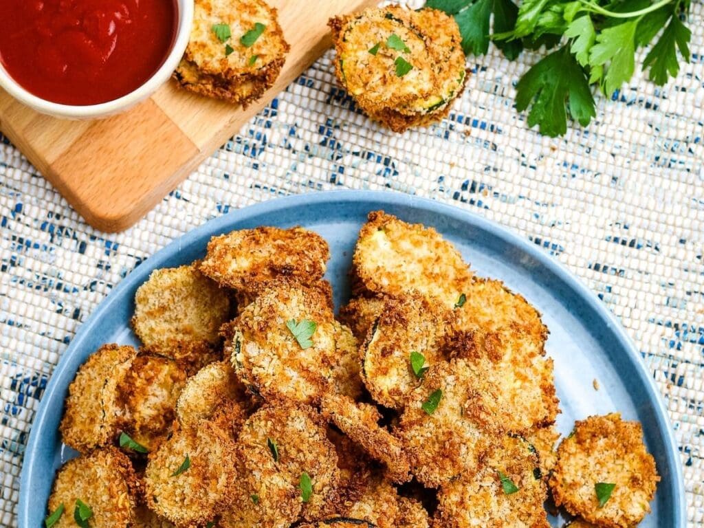 Breaded air fried zucchini chips on a blue plate with dipping sauce.
