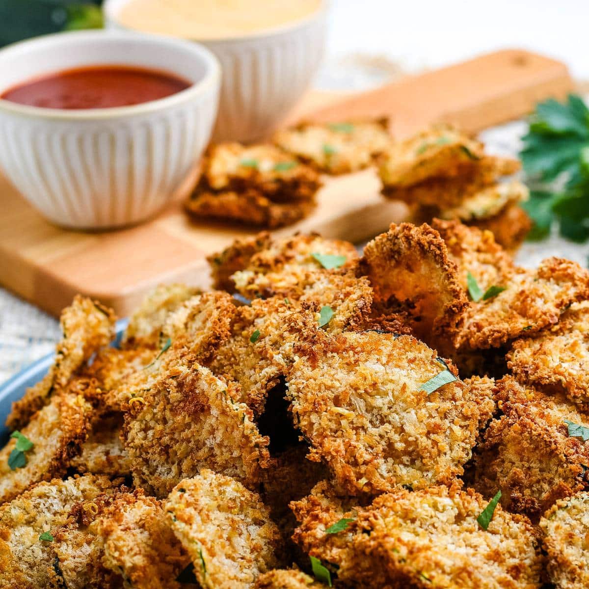 Closeup view of breaded air fryer zucchini chips with bowls of dipping sauce in the background.
