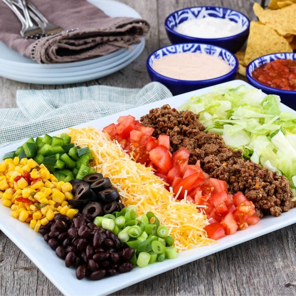 Classic taco salad ingredients on a white platter with chips and bowls of sauces in the background.