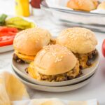 Three loose meat sliders with cheese on a stack of white plates with cheese and pickles in the background.