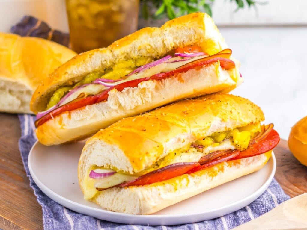 Two homemade hot Italian subs sandwiches stacked on a plate with a blue striped dish towel.