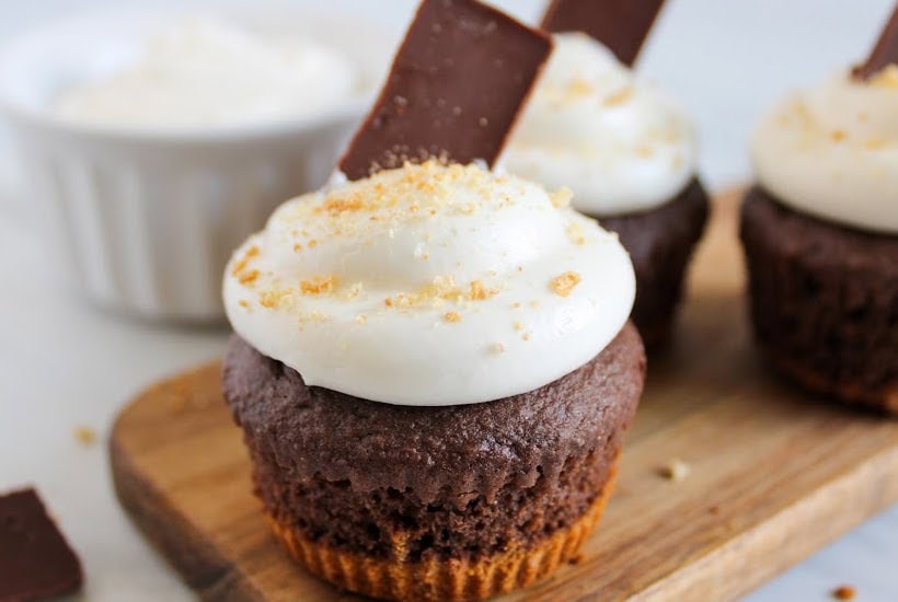 Closeup view of a smores cupcake topped with whipped marshmallow frosting and garnished with crushed graham crackers and a piece of chocolate.