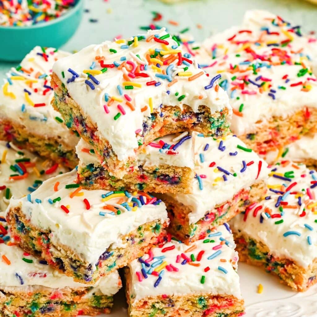 Stack of funfetti sugar cookie bars topped with white homemade frosting and rainbow sprinkles, one with a bite taken out showing the colorful funfetti sprinkles inside.