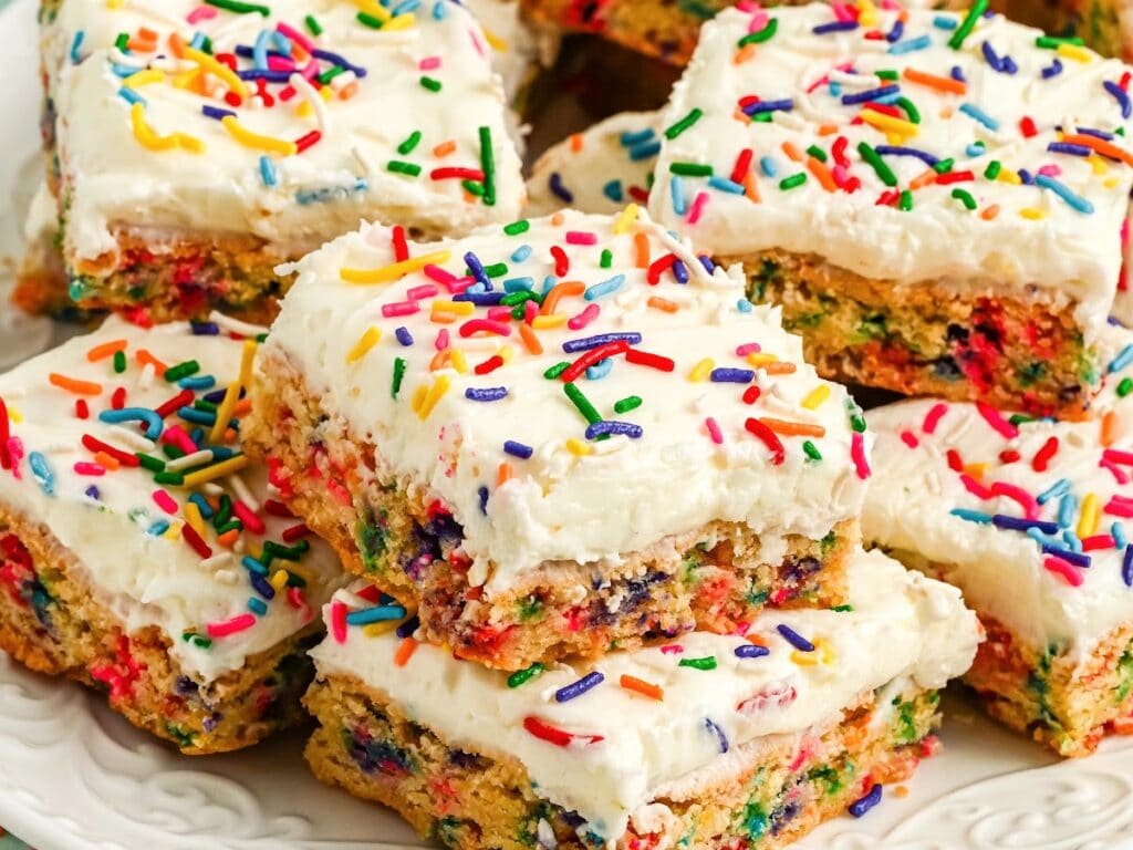 Close-up of funfetti frosted sugar cookie bars with sprinkles stacked on a white plate.