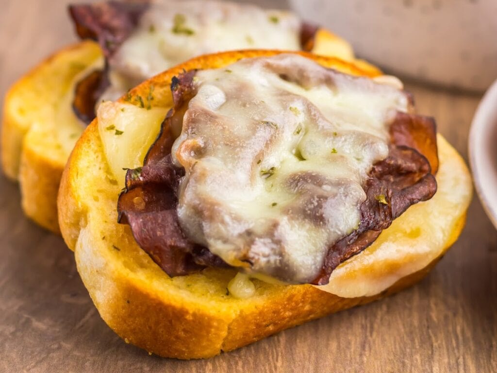 Two cheesy garlic Texas toast French dip sandwiches with provolone cheese and roast beef on a wooden board.