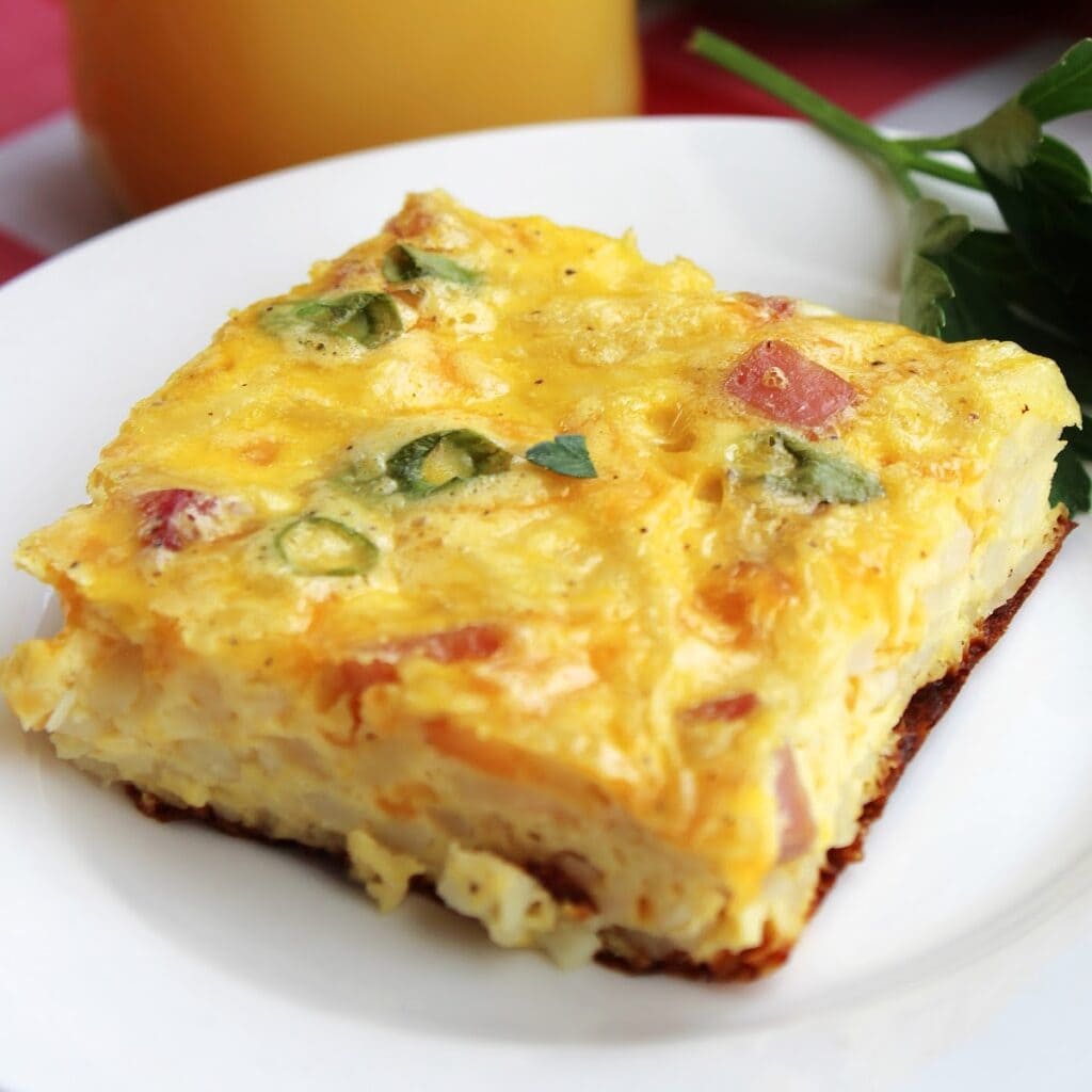 Square slice of ham egg and cheese hash brown casserole on a white plate, with a glass of orange juice in the background.