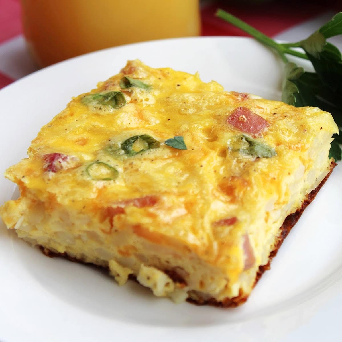Square slice of ham and cheese hash brown casserole served on a white plate, and served with orange juice in the background.
