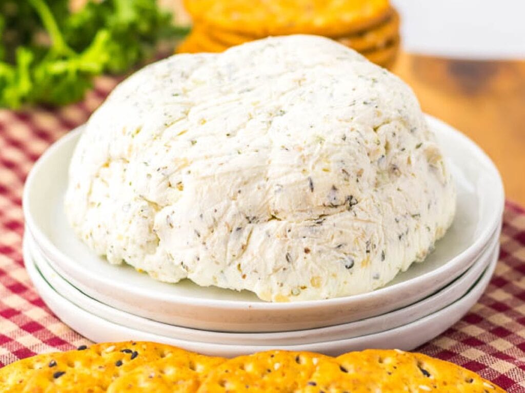 Mound of homemade Boursin garlic and herb cheese spread on a white plate, surrounded by crackers on a checkered cloth background.