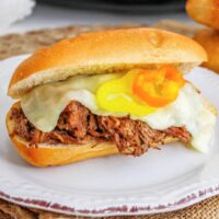 Slow cooker Italian beef sandwich on a hoagie roll topped with provolone cheese and banana peppers on a white plate.