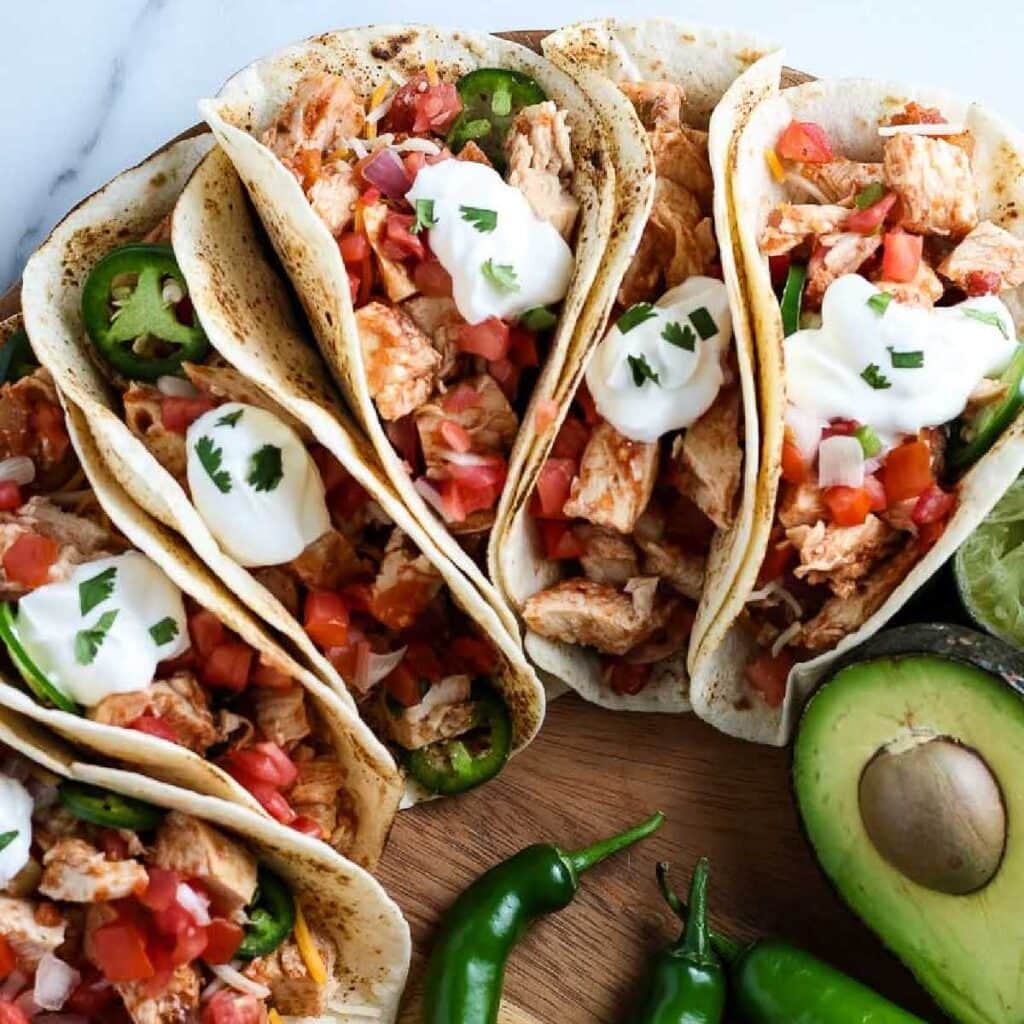 A wooden tray of crockpot salsa chicken tacos garnished with sour cream and parsley with peppers and avocado.