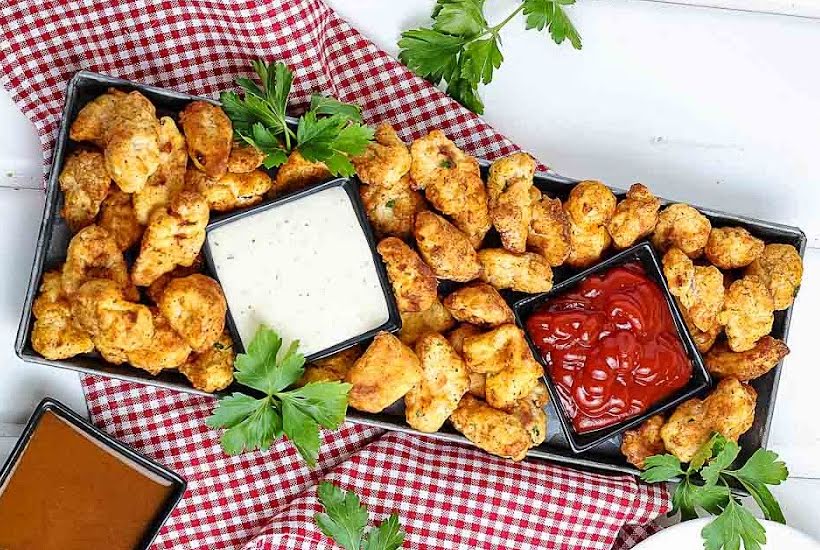 Overhead view of homemade popcorn chicken in a rectangular serving tray with a small dishes of ketchup, ranch, and bbq sauce.