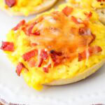 Breakfast bagel bite topped with scrambled eggs, crumbled bacon, and melted cheese on a white plate.
