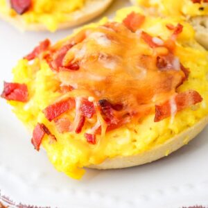 Breakfast bagel bite topped with scrambled eggs, crumbled bacon, and melted cheese on a white plate.