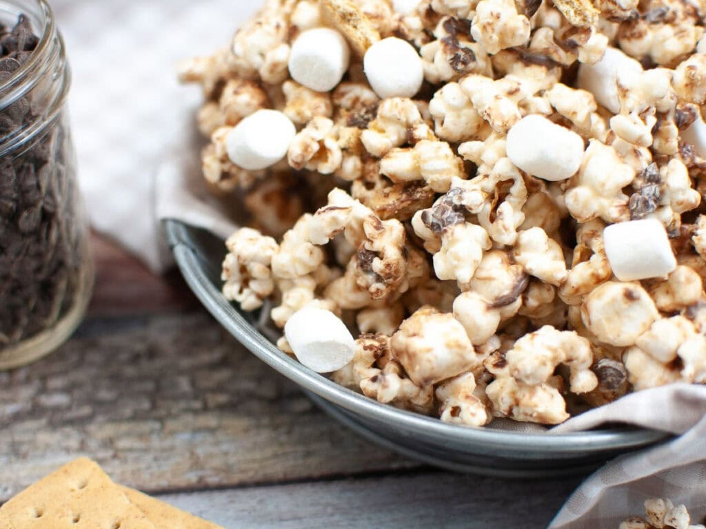 Side view of a metal bowl filled with smores popcorn with a jar of chocolate chips and graham crackers on the side.
